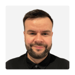 Smiling man with short dark hair and beard against a light gray background, wearing a black collared shirt. The tone is approachable and friendly.
