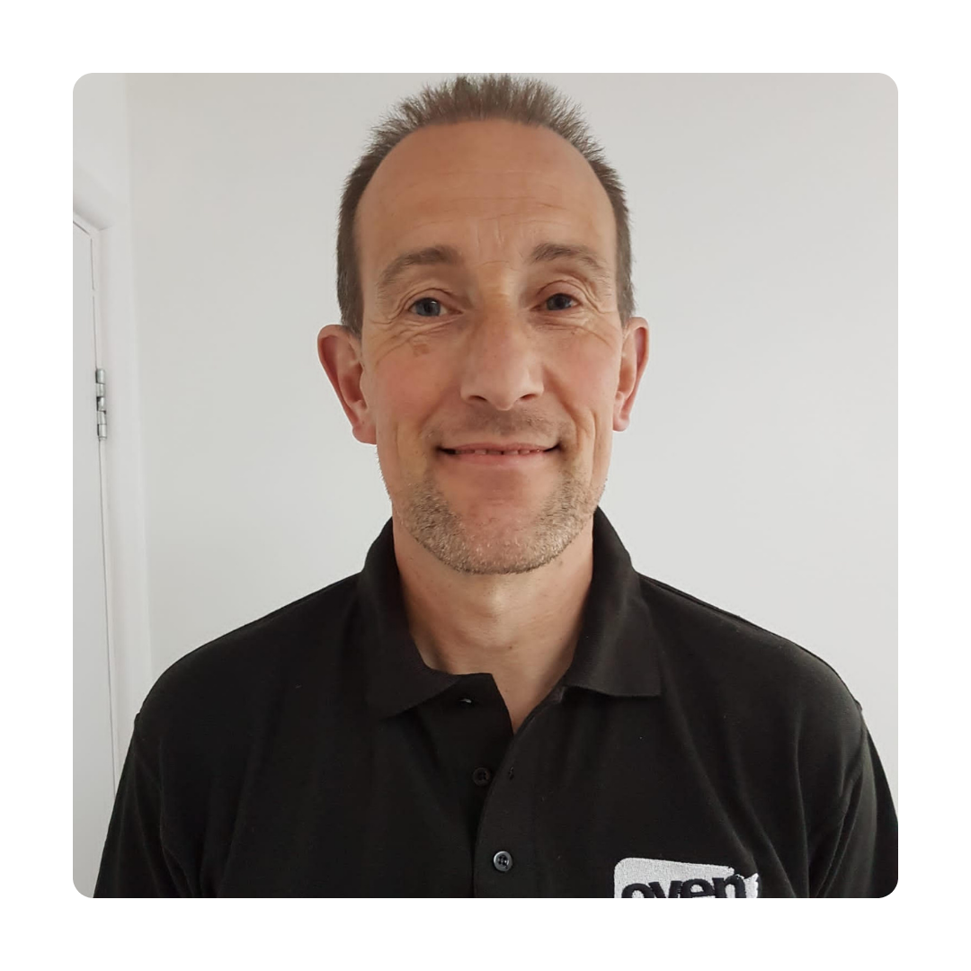 Smiling man in a black collared shirt with a logo, posing against a plain white wall. The tone is friendly and approachable.