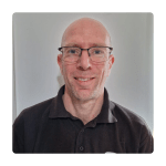Bald man wearing glasses and a black polo shirt smiles softly against a plain white background. The image conveys a friendly and approachable tone.