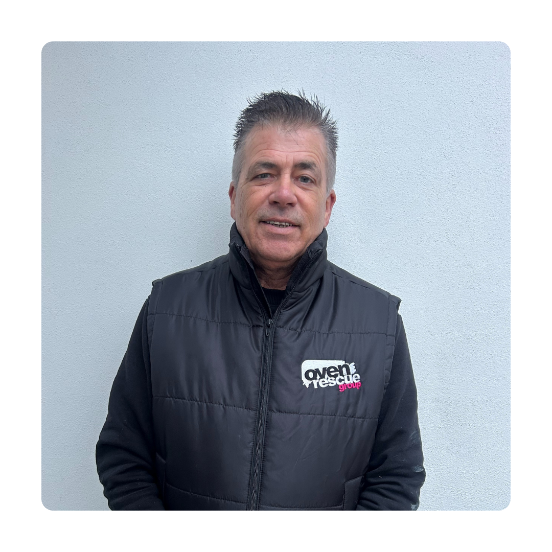 A man wearing a black vest with an "oven rescue" logo stands against a light gray wall. He is smiling, conveying a friendly and approachable demeanor.
