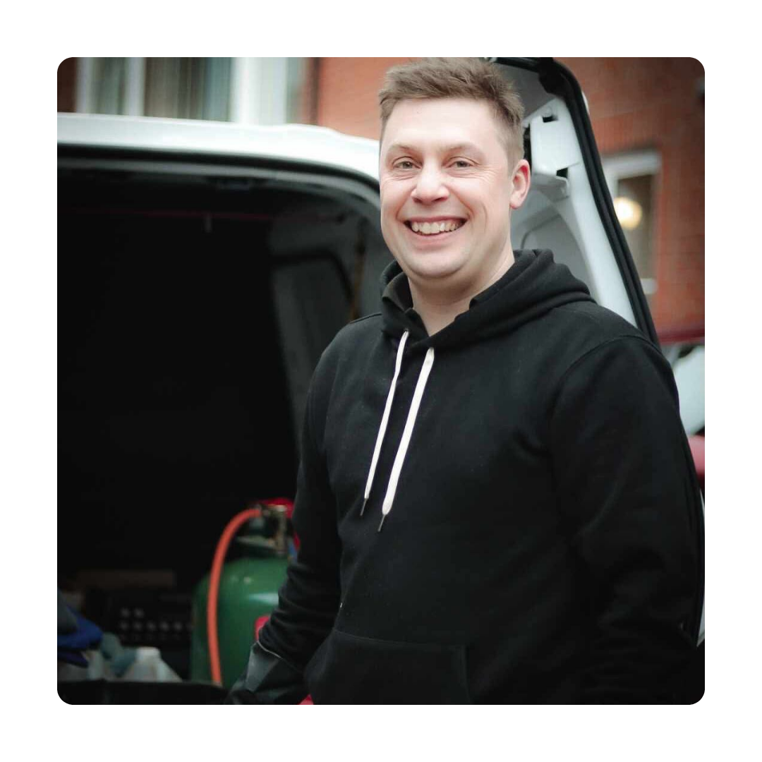 A person in a black hoodie smiles warmly while standing by an open van. The background shows a building exterior, creating a casual, cheerful scene.